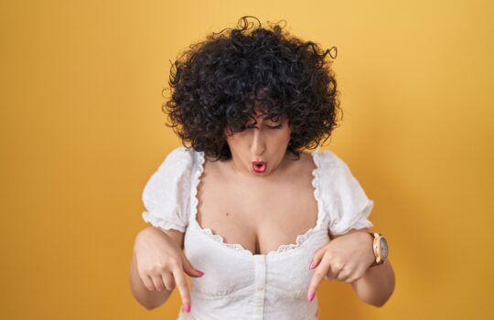 Young brunette woman with curly hair standing over yellow background pointing down with fingers showing advertisement, surprised face and open mouth