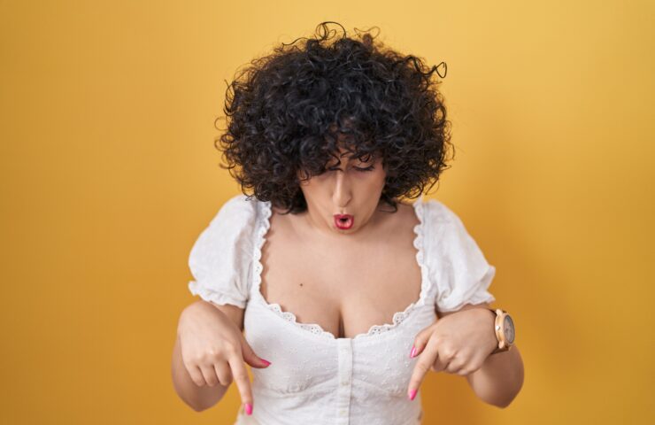 Young brunette woman with curly hair standing over yellow background pointing down with fingers showing advertisement, surprised face and open mouth